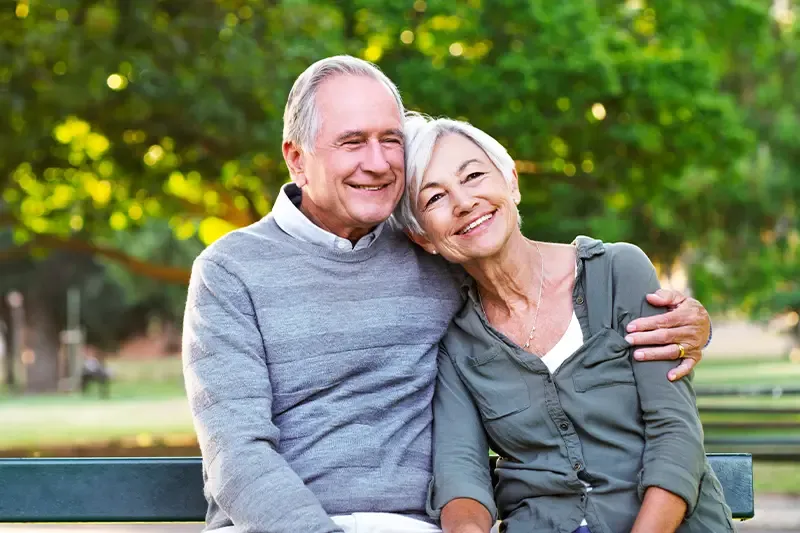 A happy elderly couple sitting on a bench in a park, with the man embracing the woman.