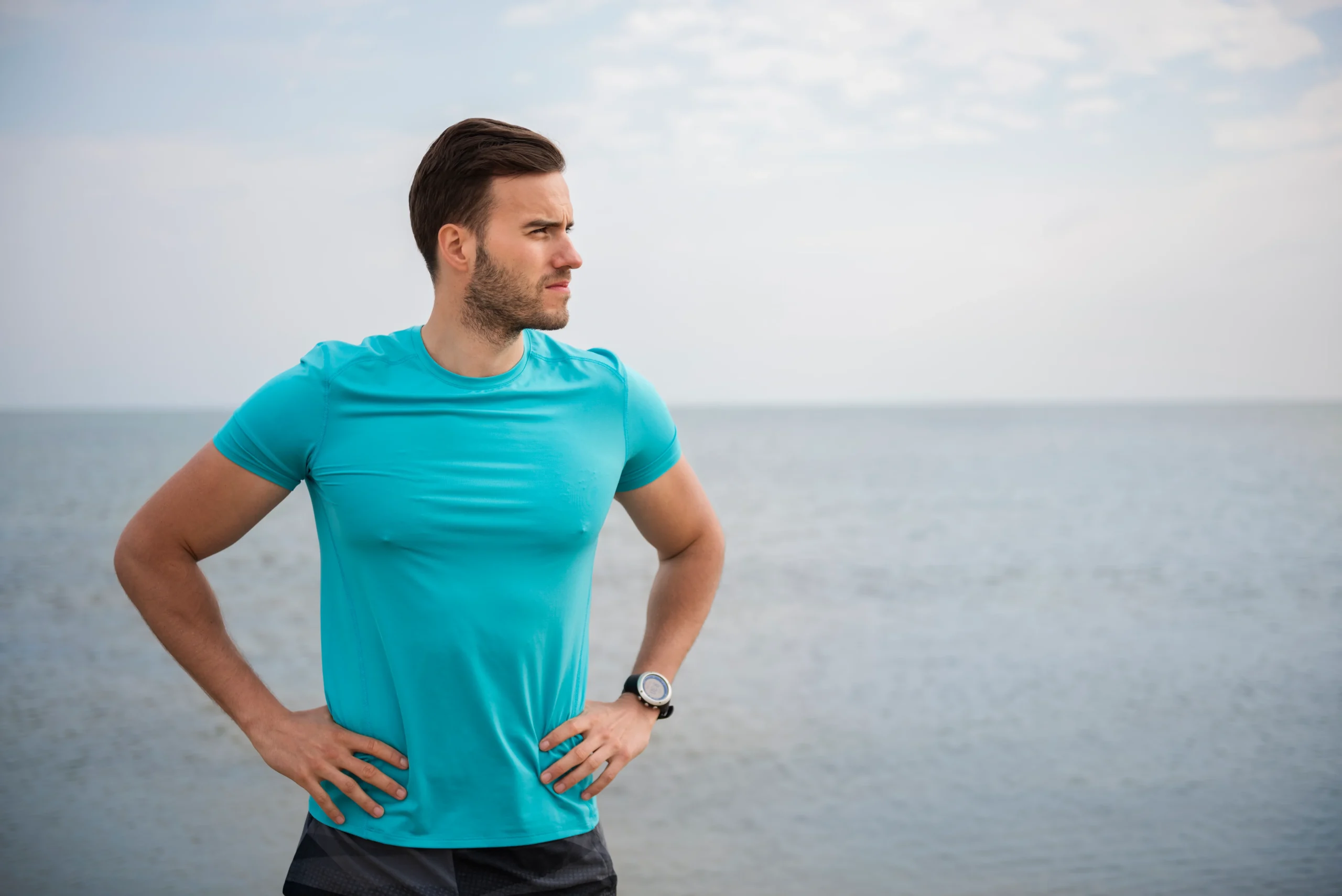 Man in turquoise athletic shirt and smartwatch standing with hands on hips by the sea under a cloudy sky.