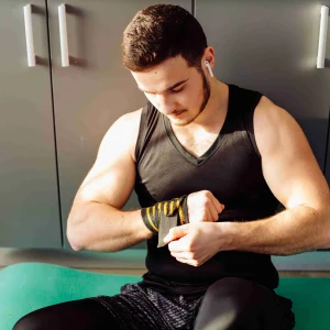 Man in black tank top sitting on a green mat, wrapping his wrist with a black and yellow strap in a home gym setting.