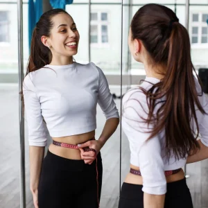 Woman in athletic wear measuring her waist with a tape measure while smiling at her reflection in a gym or fitness studio.