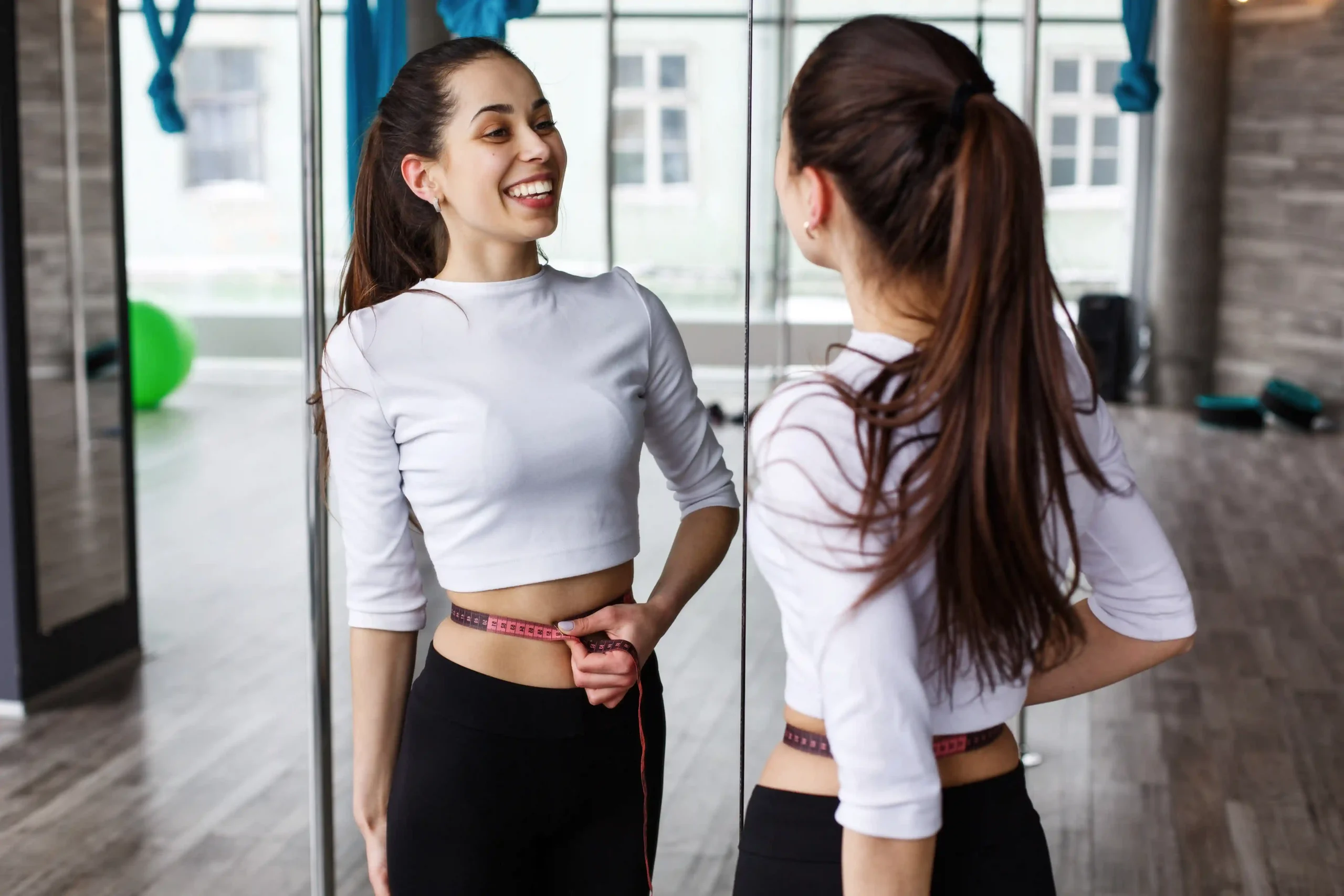 Woman in athletic wear measuring her waist with a tape measure while smiling at her reflection in a gym or fitness studio.