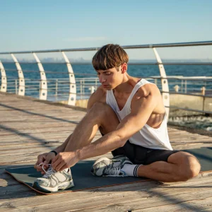 Person in athletic wear sitting on a yoga mat on a wooden pier, tying shoelaces with ocean and railing in the background.
